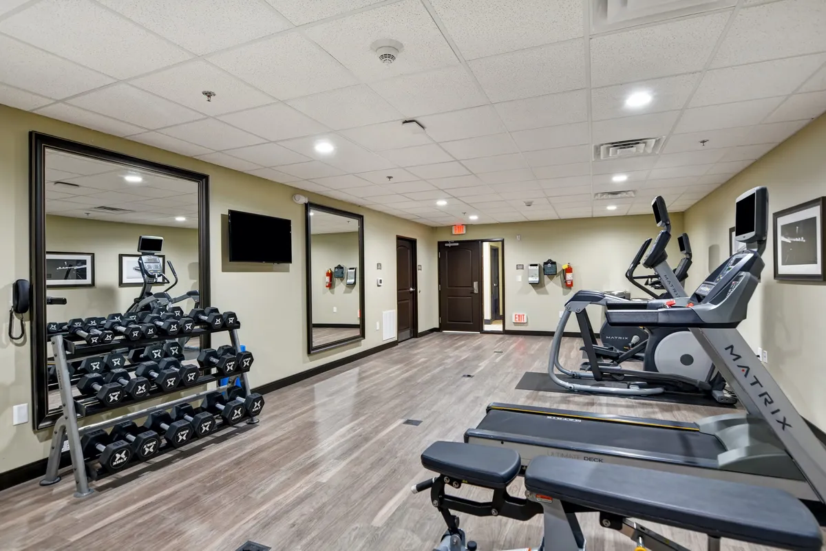 A modern fitness center equipped with a dumbbell rack, treadmills, elliptical machines, and mirrors, designed for guests to recharge after a productive day. The motivational mood encourages business travelers to maintain wellness routines while on the road.