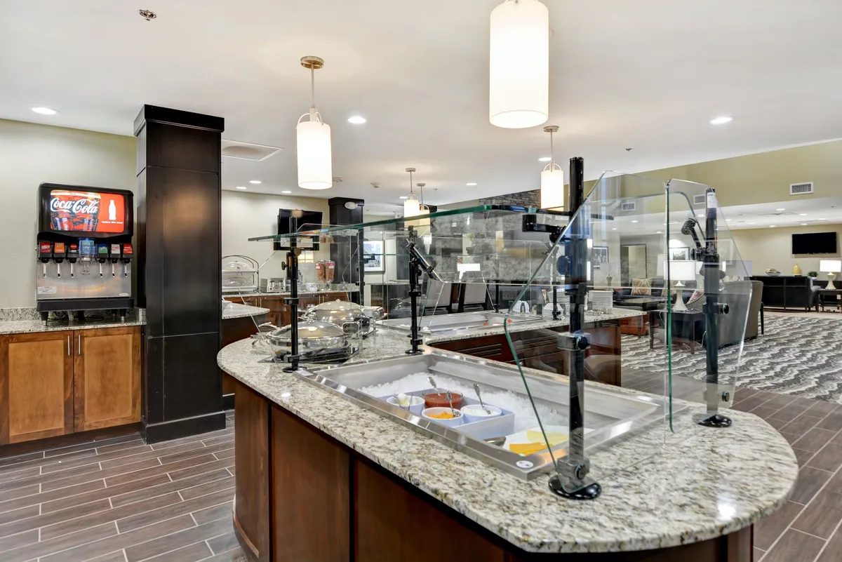This image shows a bright and welcoming breakfast area with a granite countertop displaying a self-serve food display and a beverage station. Modern lighting fixtures add to the inviting, communal atmosphere, while guests can be seen enjoying breakfast together.