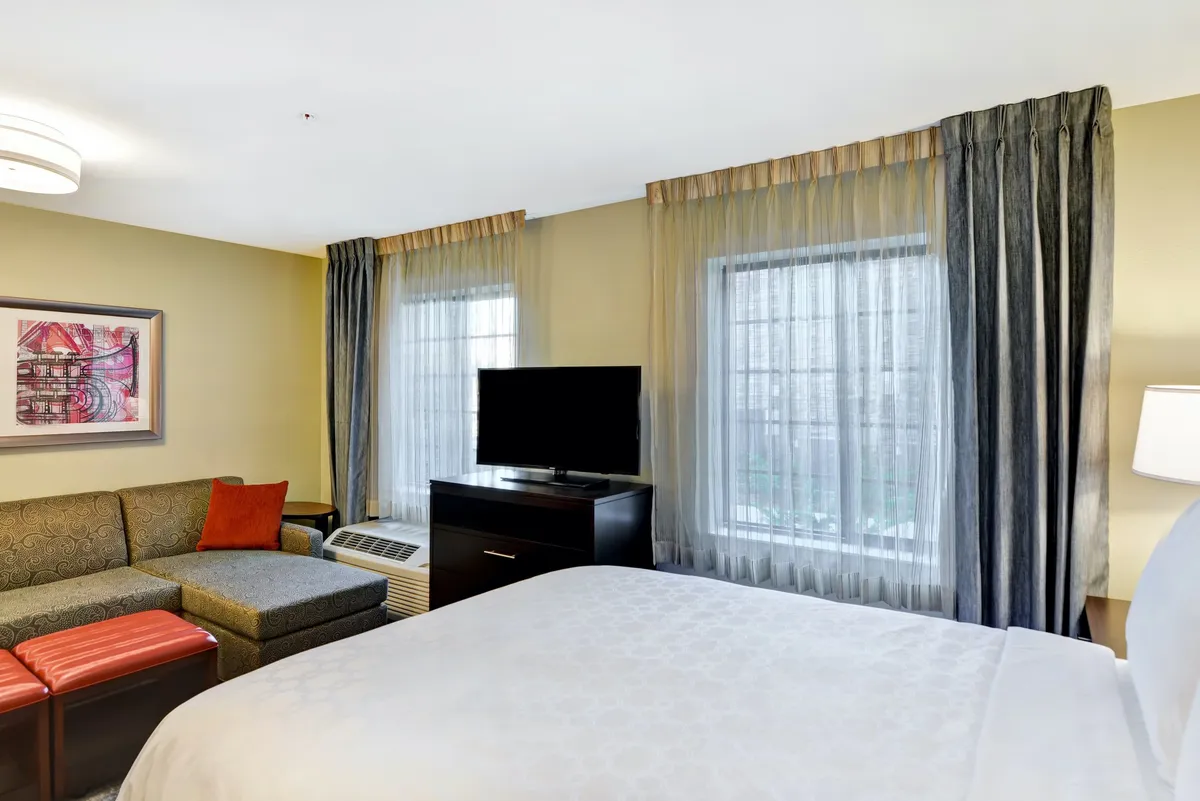 A studio suite interior featuring a queen bed with plush linens, a cozy sleeper sofa positioned for both lounging and sleeping, a wall-mounted flatscreen TV, stylish artwork, and large windows letting in natural light. The arrangement showcases an open layout combining sleeping and living areas.