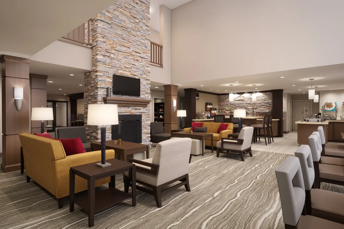 The image features a spacious, inviting lobby area with a stone fireplace, a comfortable seating area, and a reception desk in the background. The space is designed for guests to gather, socialize, or relax, with ample natural light contributing to the welcoming ambiance.