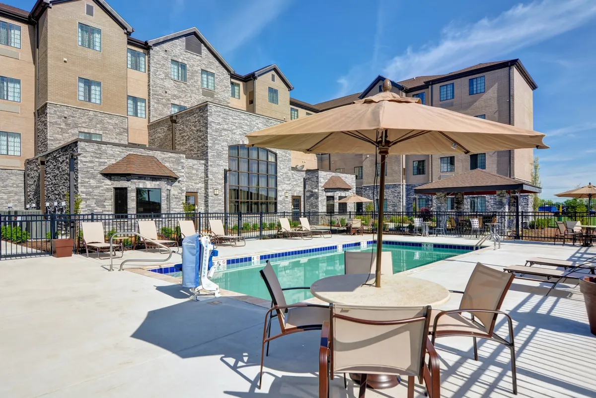 Another vibrant view of the outdoor pool, this time emphasizing the poolside seating area, umbrellas, and building facade. The mood is cheerful and leisurely, suggesting possibilities for poolside barbecues, evening swims, or sunrise yoga sessions. The image expands on the ways guests can use the outdoor space.