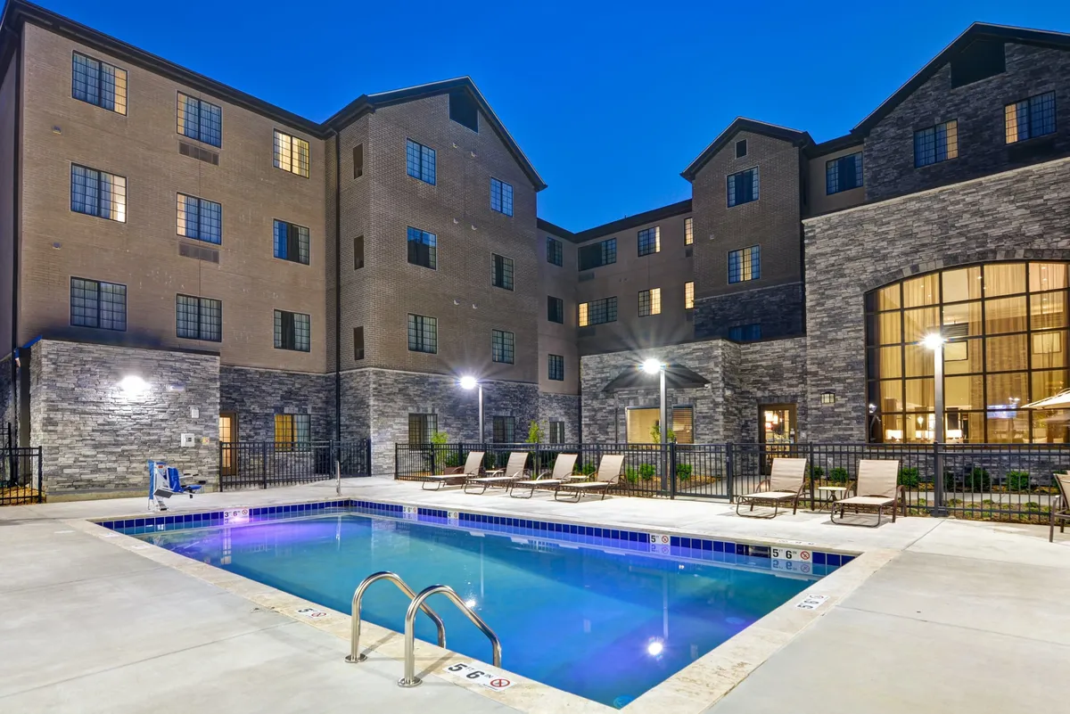 A spacious outdoor swimming pool surrounded by comfortable lounge chairs, set against a manicured landscaped area and the building facade. The scene is inviting and social, perfect for swimming, sunbathing, or gathering with friends. The area basks in sunlight, evoking a summery, relaxing vibe.