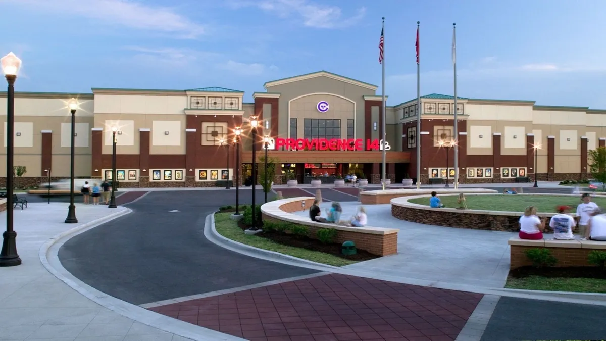 An exterior view highlighting the theater entrance at Providence Marketplace, complete with a prominent marquee sign, circular outdoor seating area, and inviting lamp posts. The setting is energetic, with people gathering to socialize and attend events.