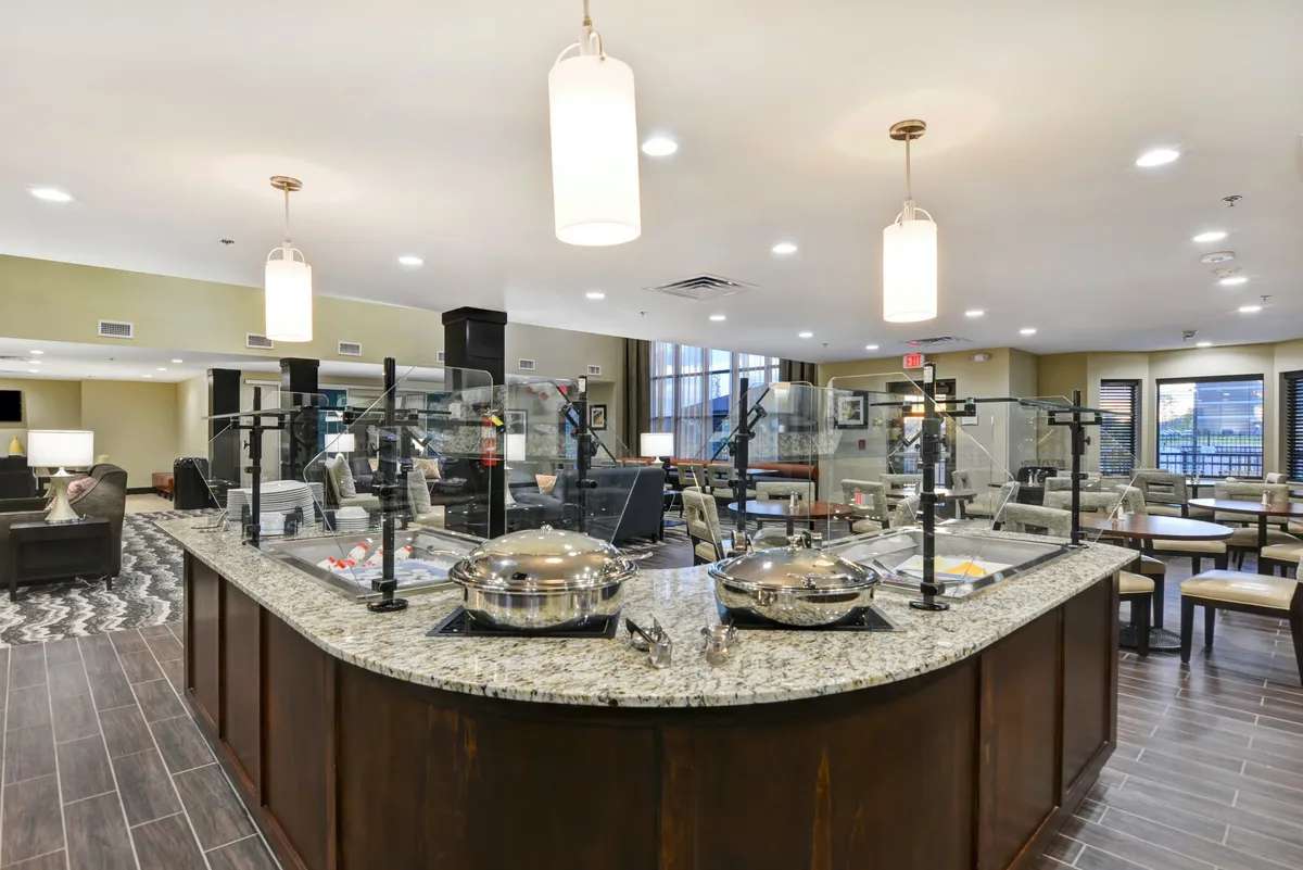 A bright and welcoming communal dining space featuring a granite countertop with food serving stations, ample seating, and large windows that fill the room with natural light. The scene suggests opportunities for breakfast, casual meetings, or quick meals before heading out.