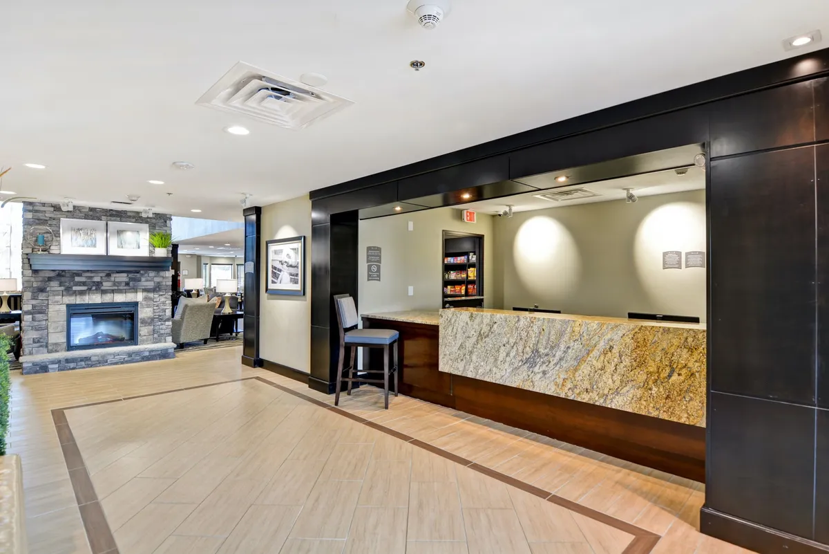 A welcoming reception area features a modern reception desk, a cozy fireplace, and comfortable seating where guests are checking in and relaxing. The inviting atmosphere showcases the property's first impression and fosters a sense of arrival and productivity for business travelers.