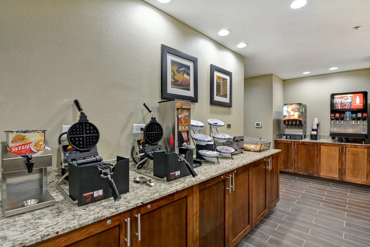 The image highlights a casual, friendly breakfast station featuring waffle makers, a syrup dispenser, a beverage station, and decorative artwork. Guests are engaged in making their own waffles and socializing, giving the space a lively, homey feel.