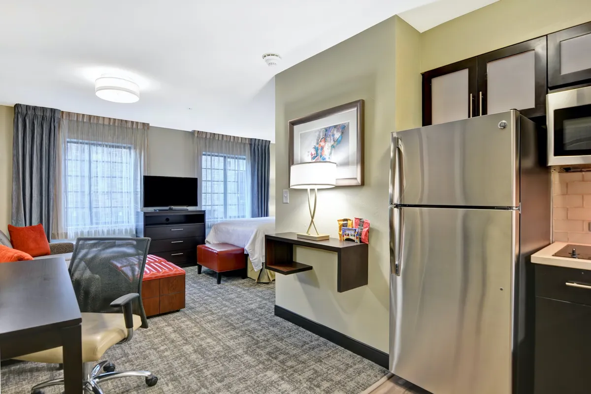 The image showcases a bright, inviting studio space featuring a modern kitchenette with a full-size refrigerator, a comfortable sofa positioned for relaxation, a dedicated desk area for work or planning adventures, and large windows that fill the room with natural light. The setup highlights both functionality and comfort, perfect for a couple's getaway or a solo retreat.
