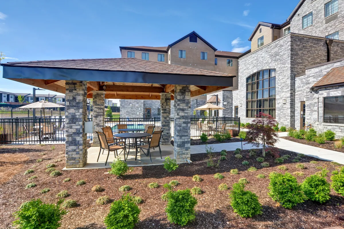 This image showcases an attractive stone gazebo surrounded by a landscaped garden. There's a dining table and chairs set up under the gazebo, and the scene radiates a peaceful, sunlit atmosphere perfect for outdoor relaxation, dining, or social gatherings. Hints of a pool area add to the ambiance of leisure and connection with nature.