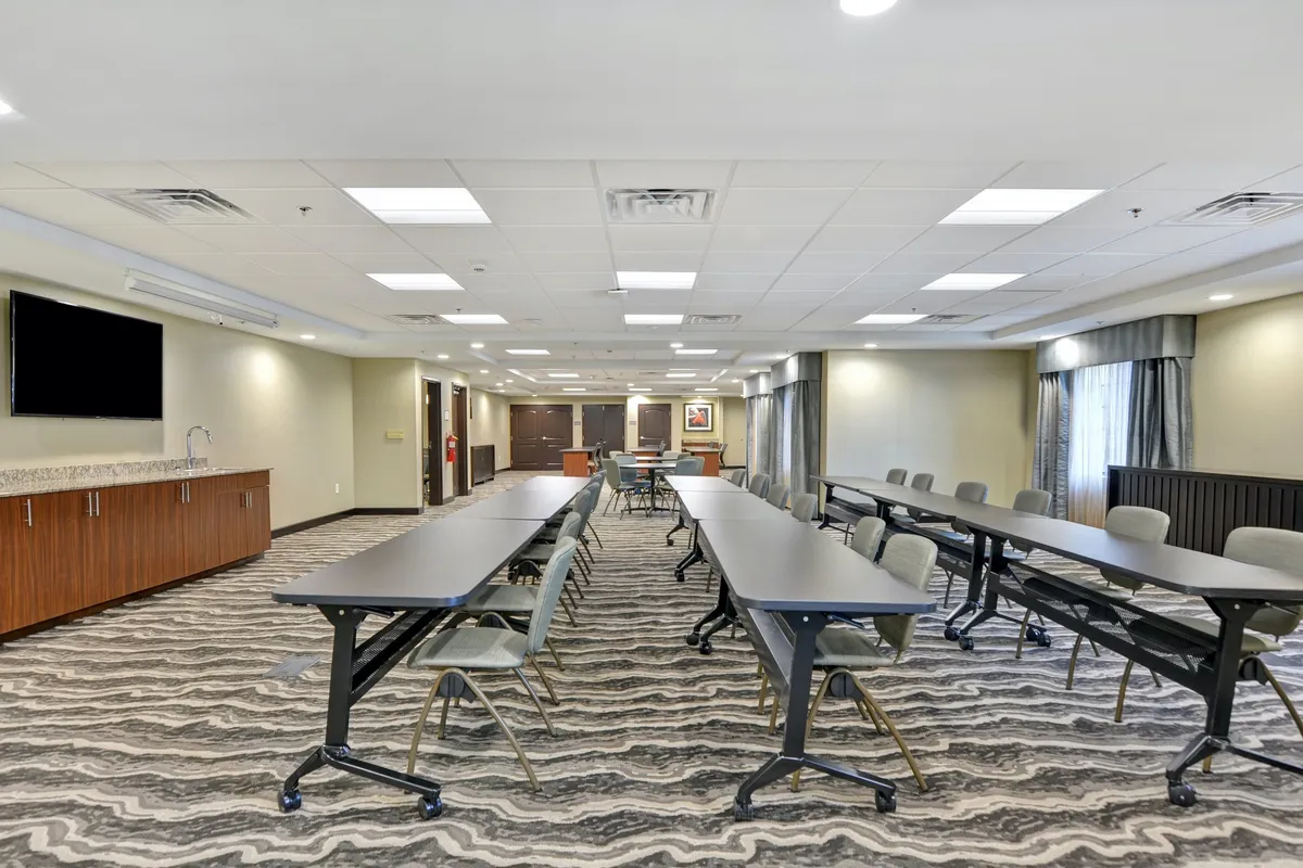 A modern business center interior with long tables arranged for meetings, a large television screen mounted on the wall, and sleek wooden cabinetry, creating a collaborative and focused environment for work. The space is well-lit and designed to support productivity, with ample room for laptops, notepads, and small group discussions.
